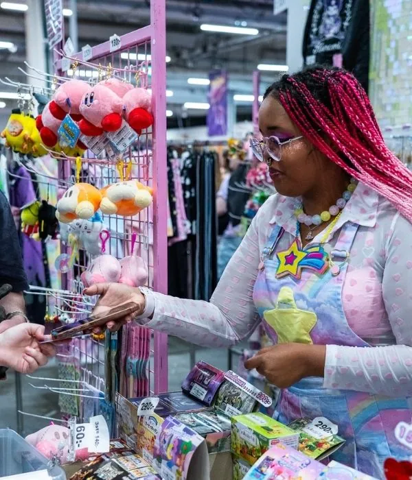 A vendor with bright pink braids serving a customer at a plush toy and collectibles stall